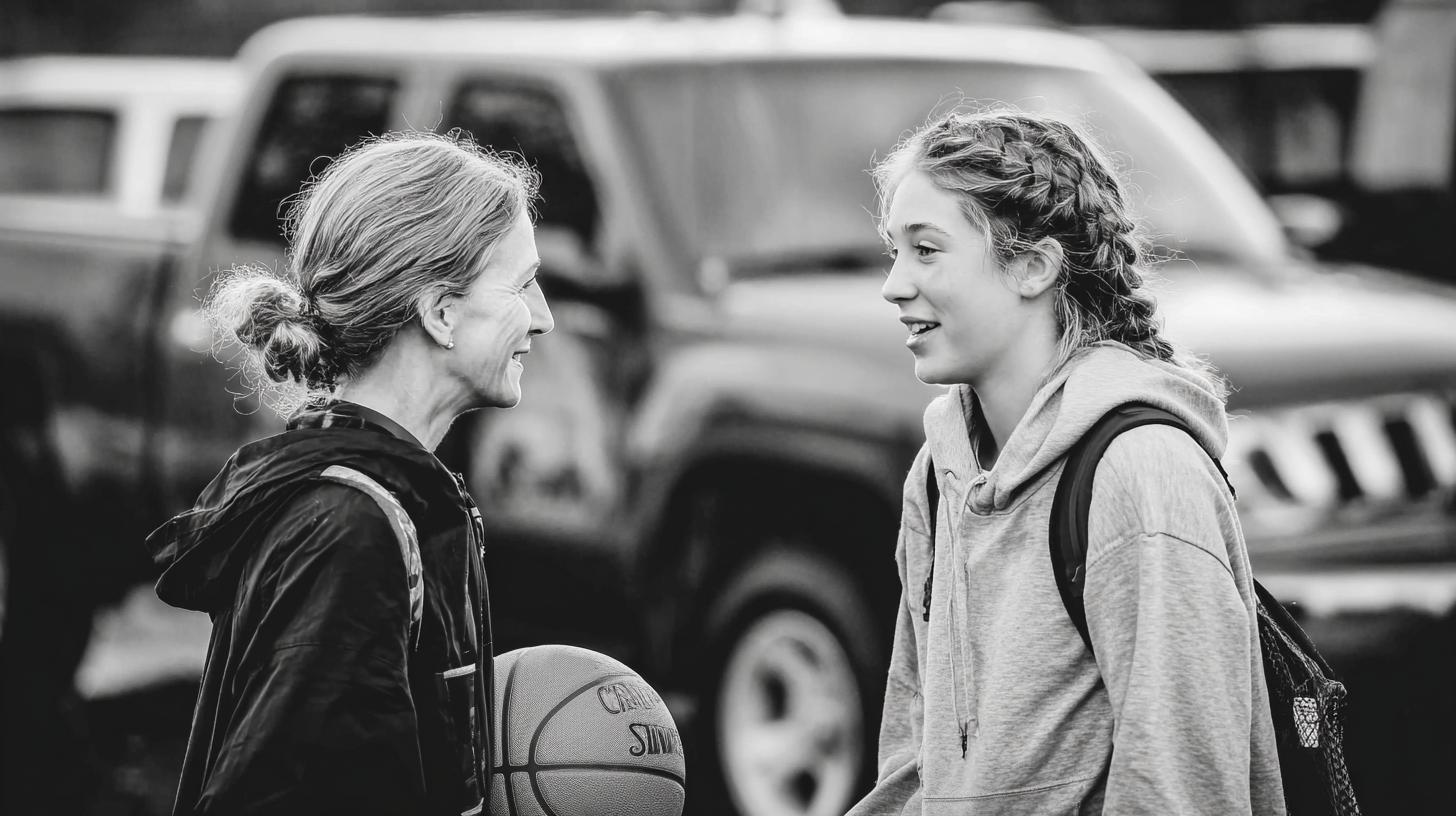 Two women, one with a basketball, standing next to each other outdoors with vehicles in the background.